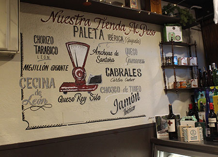 Un cartel pintado en la pared dónde podemos ver una báscula tradicional y los diversos productos que se pueden adquirir: Paleta ibérica (Guijuelo), chorizo tarabico (León), anchoas de Santoña, Queso Gamoneu, Cabrales...