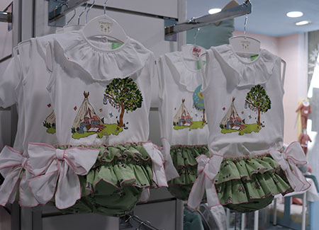 Vista del interior del comercio, dónde vemos expuestas prendas infantiles para niña de color blanco y verde con estampados de una tienda de campaña en la naturaleza