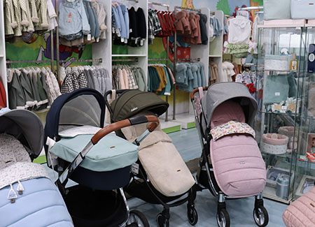 Vista del interior del comercio, dónde vemos tres sillitas de bebé, de diferentes estilos y colores, junto a un carrito de bebé. Al fondo vemos varios expositores con barras llenas de ropa colgada de perchas