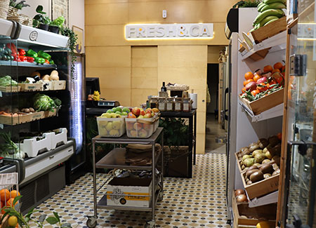 Vista del interior del comercio, dónde podemos ver el mostrador al fondo, las estanterías con fruta y verduras contra las paredes y un expositor en el centro con cestas de fruta