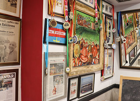 Vista de una pared dónde podemos ver marcos con recortes de periódicos, medallas, diplomas, un cuadro y una bandera de Asturias