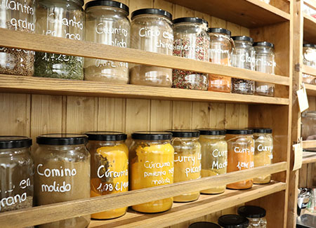 Una estantería de madera con varios tarros de cristal rellenos con especias de diversos tipos: Comino molido, Cúrcuma, Curry, Pimienta...