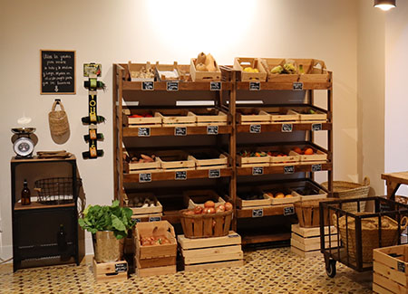 Vista del interior de la tienda dónde se puede ver una estantería doble de madera con fruta y verduras, frente a ellas hay unas cestas y cajas con más productos.