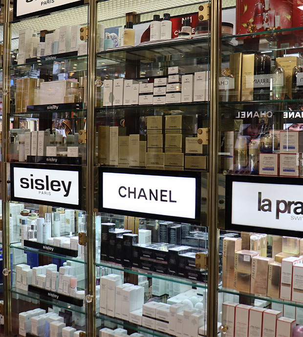 Vista del interior de la perfumería, dónde podemos ver una estantería de metal y cristal cerrada que contiene múltiples productos. También vemos carteles con las marcas: Sisley, Channel y La Prada