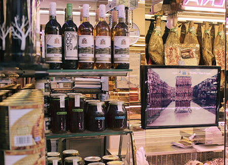 Vista del interior de la carnicería dónde se pueden ver botellas de vino, tarros de miel, botes de conservas, una esquina de un mostrador expositor y, al fondo, parte de la selección de jamonos