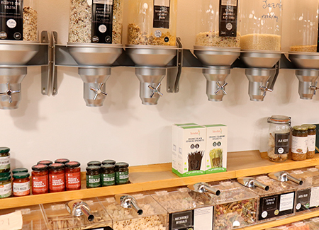 Vista de una pared con dispensadores de productos a granel por gravedad y una estantería con expositores y tarros de productos artesanos