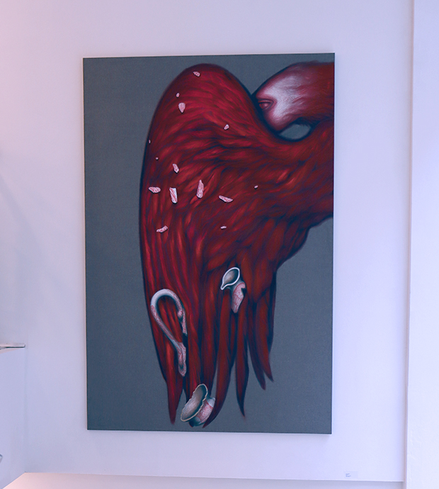 Primer plano de una de las obras expuestas, dónde se puede ver lo que parece el ala de un pájaro de color rojo con varios objetos sobre ella, y con su cabeza escondiéndose por detrás de ella