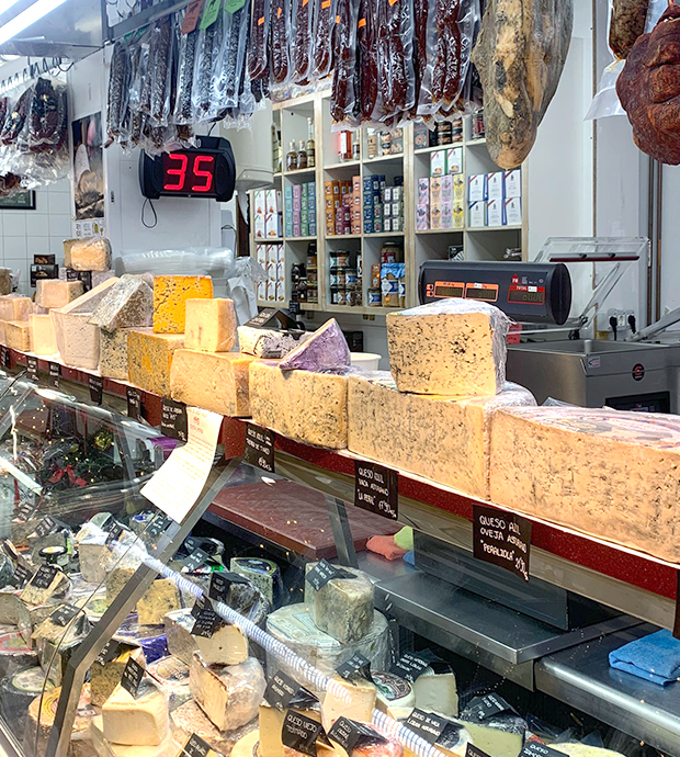 Vista del mostrador expositor del comercio, en el que vemos una selección de quesos de diversos tipos. Sobre el mostrador, vemos colgados diversos embutidos y jamones