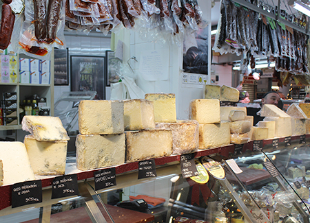 Vista del mostrador expositor del comercio, en el que vemos una gran selección de quesos. También podemos ver embutidos colgados