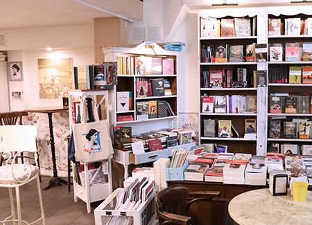 Vista del interior de la librería, dónde podemos ver estanterías y expositores con libros y bolsas de tela de la librería, junto a mesas y sillas de la zona de la cafetería
