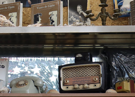 Primer plano de una balda dónde se ve una radio antigua, un teléfono antiguo de disco, un transformador de 220v a 125v. Sobre esta balda hay otra con CDs, caracolas, un candelabro y sobre la pared del fondo fotos de artistas