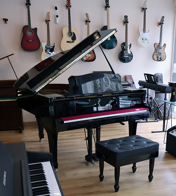 Vista del interior del comercio, dónde podemos ver un piano de cola con su taburete, en primer plano y al fondo podemos ver teclados electrónicos y en la pared del fondo vemos una colección de guitarras eléctricas y acústicas