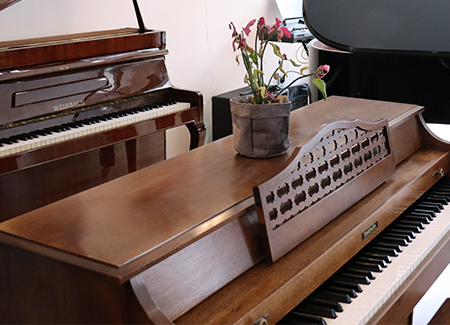 Primer plano de un piano de color madera, detrás de el se ve un piano de cuerpo vertical y la cola de un piano de cola negro