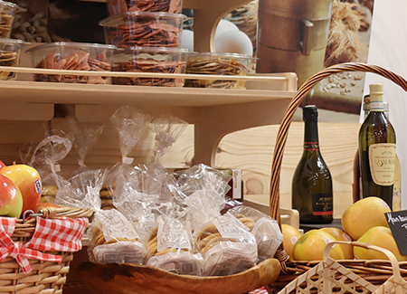 Primer plano de cestas de fruta, junto a una cesta con productos dulces artesanos, al fondo se ve un estante con más productos y unas botellas de vino