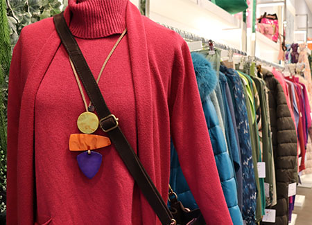 Un perchero con varias prendas de abrigo colgadas en perchas y, en primer plano, un maniquí con un jersey y una chaqueta de color rojo, un colgante de tres piezas de grandes dimensiones y un bolso cruzado