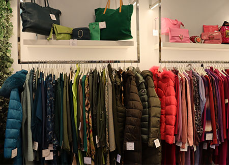 Un perchero de pie con múltiples prendas de abrigo en perchas y, al fondo, dos estanterías de pared con bolsos